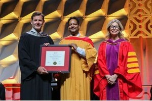 Kristian Bainey receiving a framed award on stage from academic officials in regalia